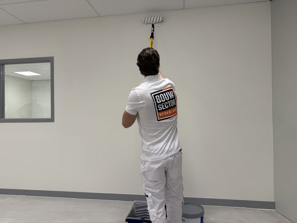 Schilder in wit tenue rolt het bovenste deel van een lege wand, met verfbak en emmer aan zijn voeten