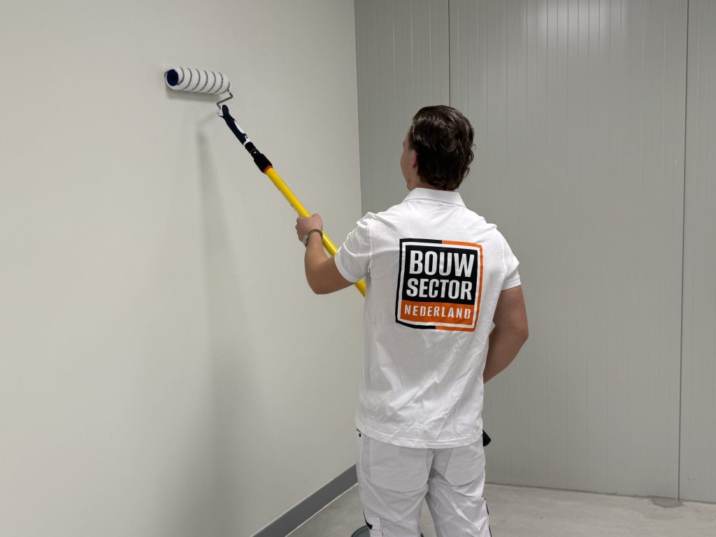 Schilder in witte kleding zet een roller met lange steel op een witte wand