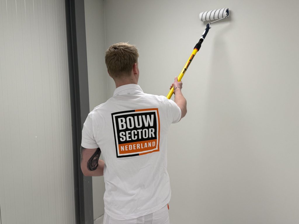 Schilder met het logo van Bouwsector Nederland op zijn rug rolt een wand in een smalle gang