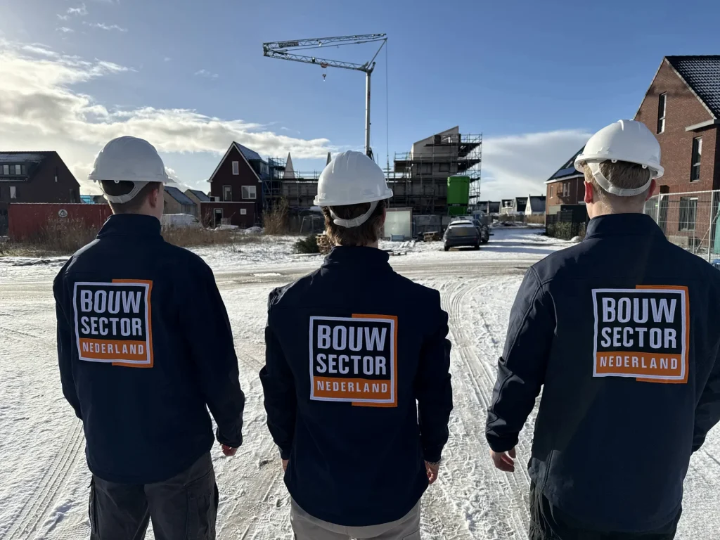 Drie vakmensen in donkerblauwe jassen met logo kijken uit over een woonwijk in aanbouw