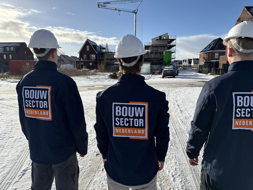 Drie werkjassen met het logo van Bouwsector Nederland op een besneeuwde bouwplaats bij een rode woning