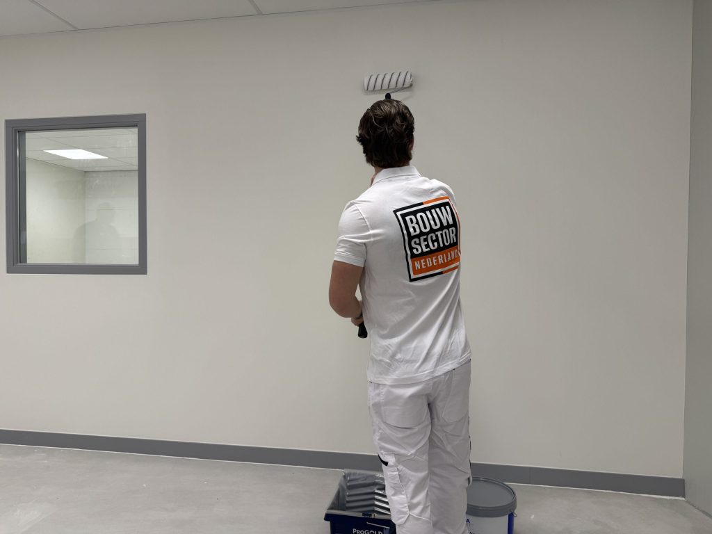 Schilder van Bouwsector Nederland rolt een witte wand, met verfbak en emmer aan zijn voeten