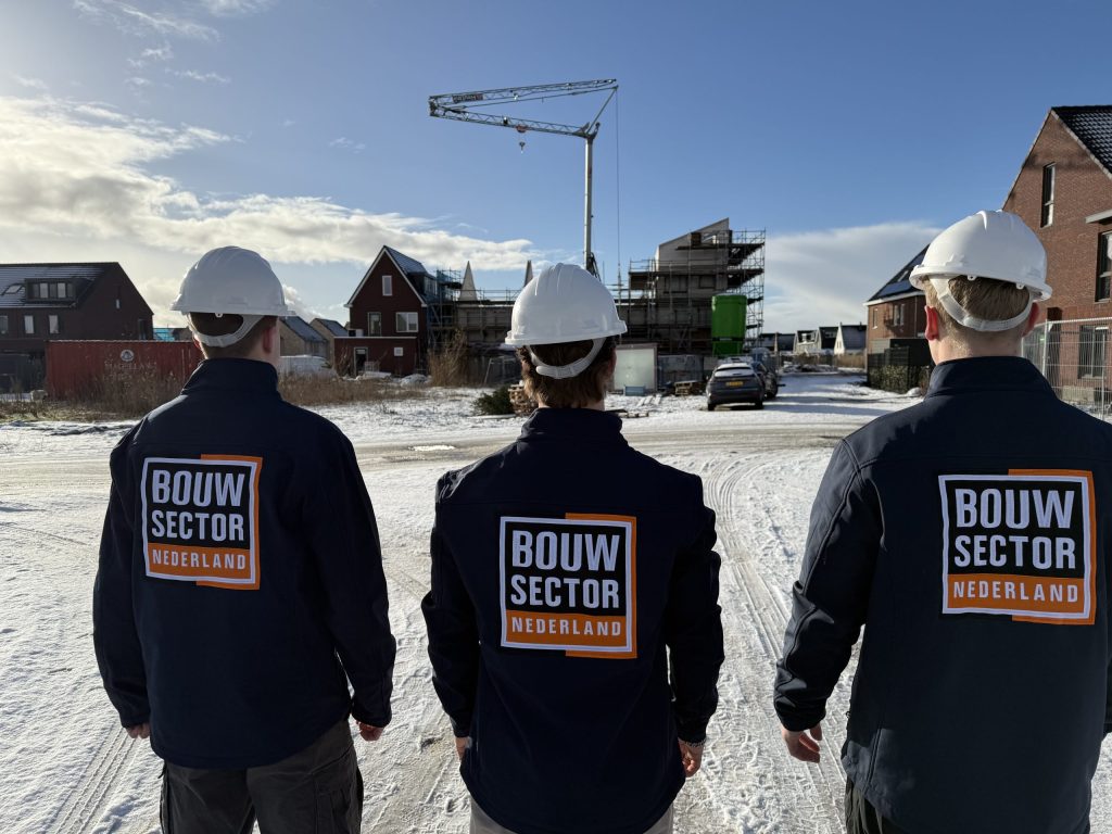Drie vakmensen van Bouwsector Nederland in werkjas met logo bij nieuwbouwwoningen en een bouwkraan